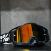 A pair of black high-performance motocross goggles with orange tinted lenses resting on a dark surface.