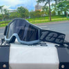 Gray off-road motorcycle goggles resting on a silver surface with a blurred green background.