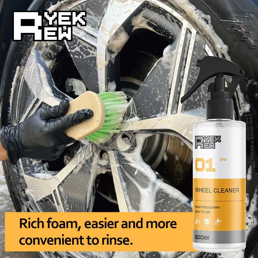 A hand is using a green brush to clean a wheel with yellow liquid foam from a bottle of wheel cleaner.