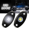 Pair of black LED rock lights with yellow illumination shown next to a blue Jeep.